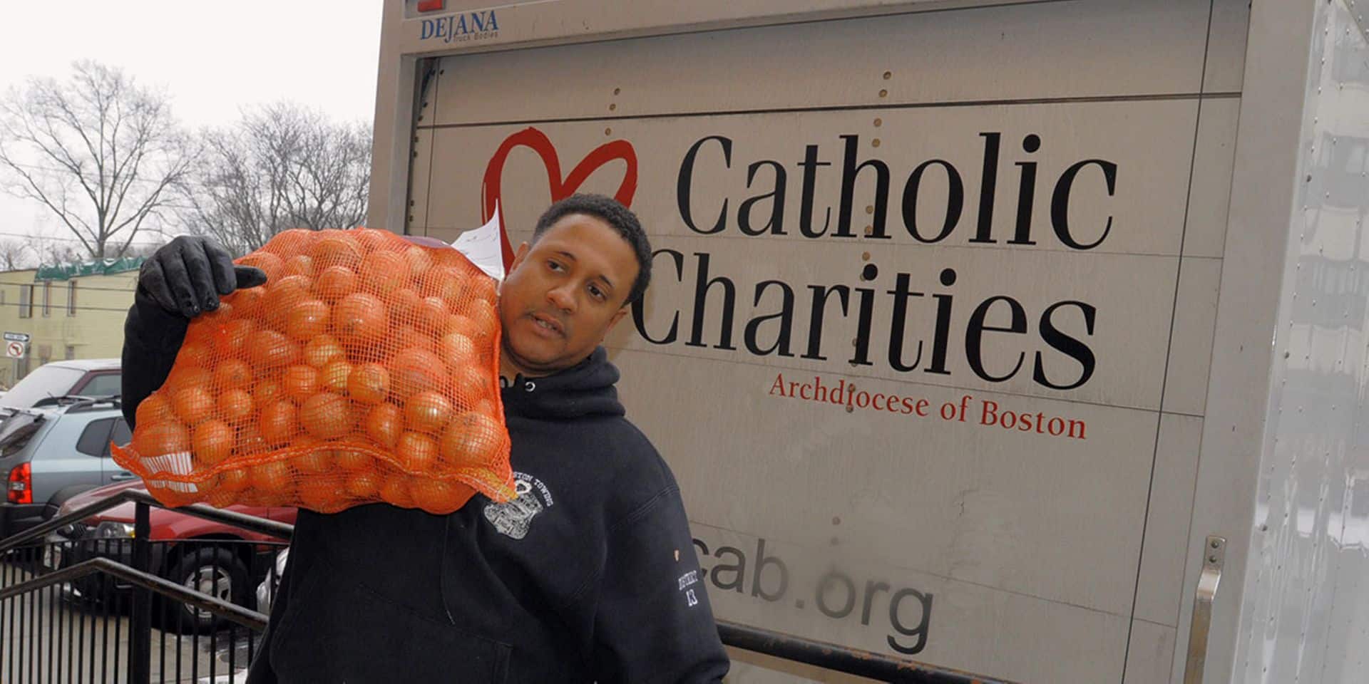 Person holding bag of oranges by Catholic Charities truck, community aid.